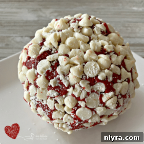 A finished Red Velvet Cheese Ball, round and covered in white chocolate chips, presented on a dark background.