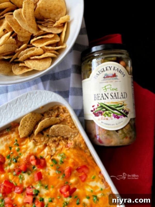 A warm, cheesy Hot Taco Five Bean Dip served in a baking dish, with a scoop taken out and surrounded by tortilla chips, ready for sharing.