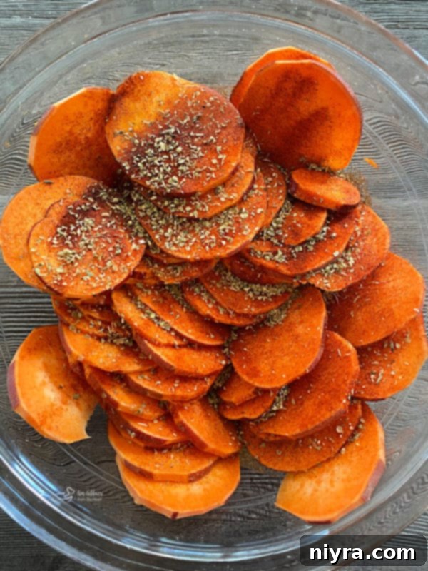 Sweet potato slices in a bowl, sprinkled generously with paprika, seasoned salt, and oregano, ready to be tossed.