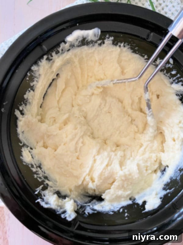 Creamy, mashed potatoes in the Crockpot with a potato masher, showing smooth texture.