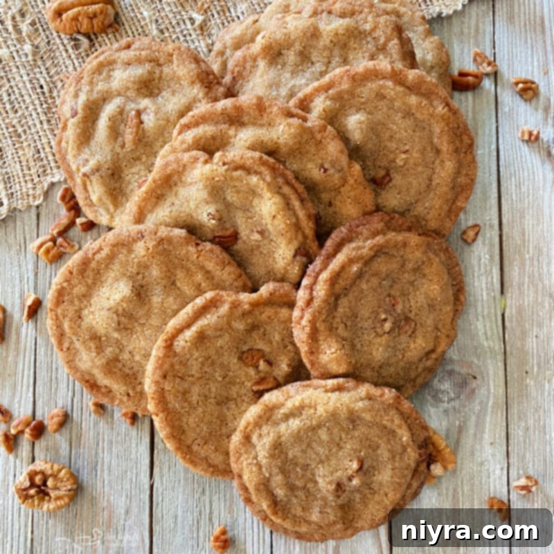 A close-up shot of several pecan butter cookies, glistening with a slight sugar crust, arranged appealingly on a light-colored surface, with whole pecans visible in the background.