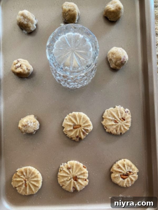 Raw pecan butter cookie dough balls, carefully spaced on an ungreased baking sheet, adorned with decorative fork imprints, ready for the oven, with a glass and whole pecans nearby.
