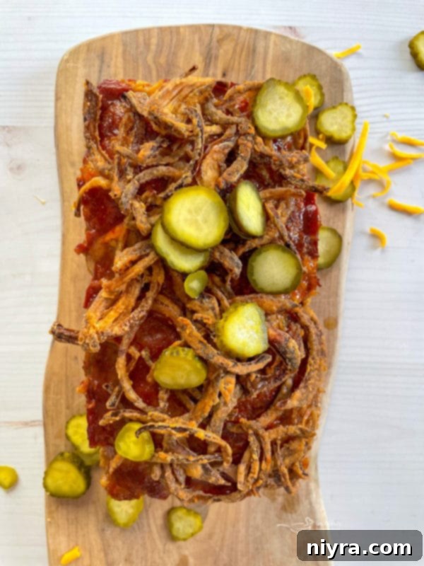 A perfectly baked Bacon Cheeseburger Meatloaf, freshly removed from the oven and topped with a generous layer of crispy fried onions and vibrant green pickles, ready to be sliced and served.