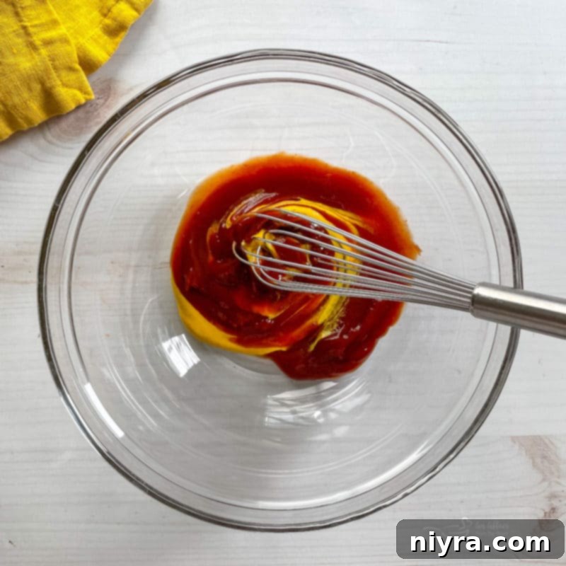 A whisk blending ketchup and mustard in a small white bowl, creating a smooth, tangy sauce for the meatloaf.