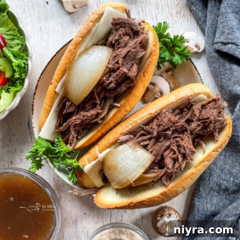 A close-up of two French Onion French Dip sandwiches, with tender beef, melted provolone, and visible caramelized onions peeking out, accompanied by a small bowl of savory au jus.