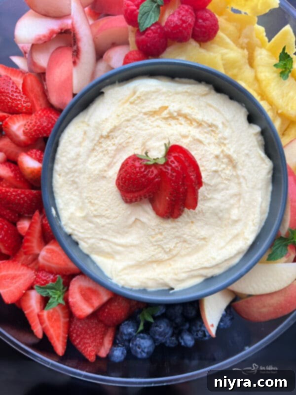 A bowl of creamy vanilla almond fruit dip positioned on a white ceramic plate, surrounded by vibrant fresh fruits like raspberries, blueberries, grapes, and strawberries.