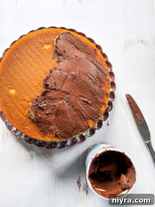 A yellow cake layer being generously frosted with rich chocolate icing, creating a smooth surface for the Kit Kat bars.