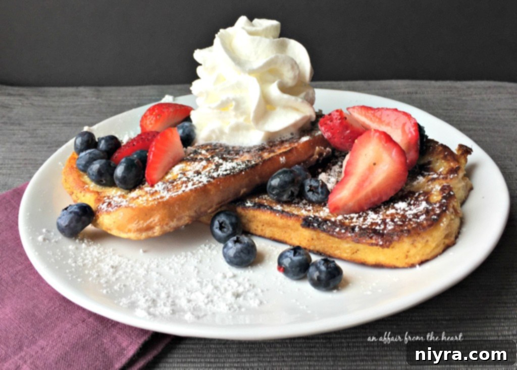 Two pieces of French Toast on a white plate, served with berries and powdered sugar, and maple syrup.
