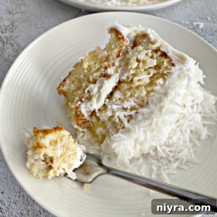 A fork with a bite of the COCONUT CREAM POKE CAKE WITH COCONUT WHIPPED CREAM FROSTING next to the slice of cake on a white plate