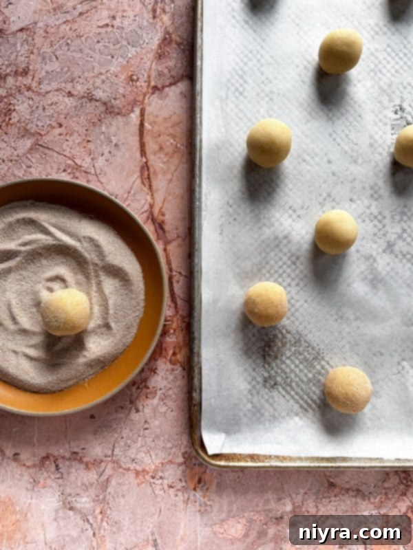 Rolling balls of snickerdoodle dough in a cinnamon-sugar mixture before shaping into thumbprints.
