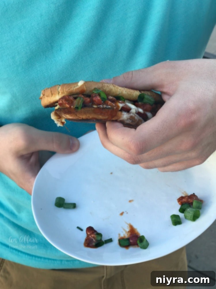 A hand holding a finished Spicy Grilled Cheese Chili Dog, showcasing the rich chili and vibrant green onion toppings, ready to eat.