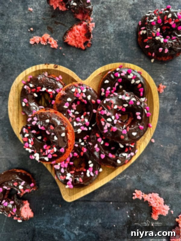 Two baked chocolate covered strawberry donuts stacked on a white plate, garnished with fresh strawberries and sprinkles.