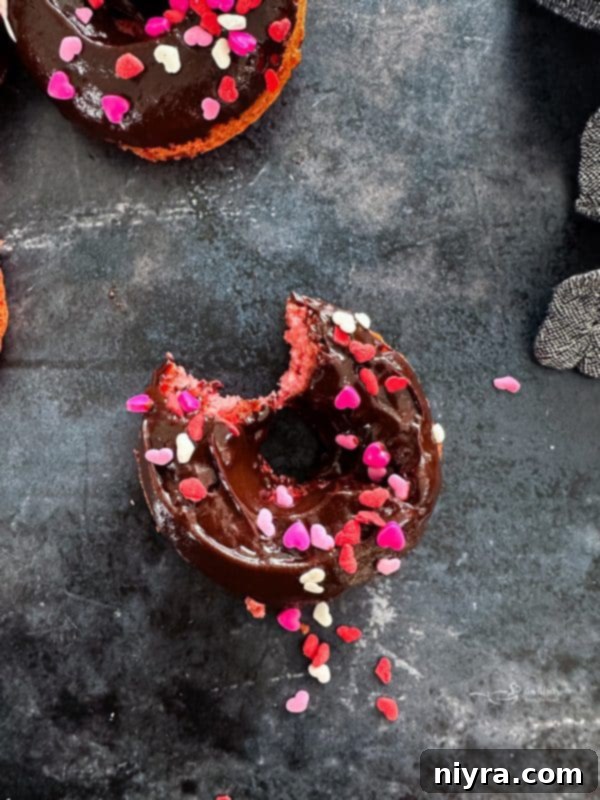 A close-up of a stack of baked chocolate covered strawberry donuts with sprinkles on a white background.