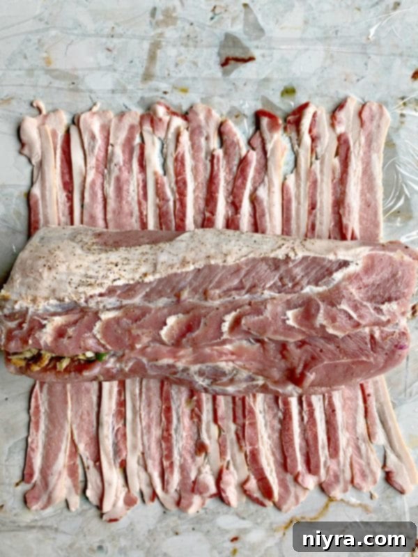 Pork tenderloin being wrapped with thick-sliced bacon on plastic wrap.