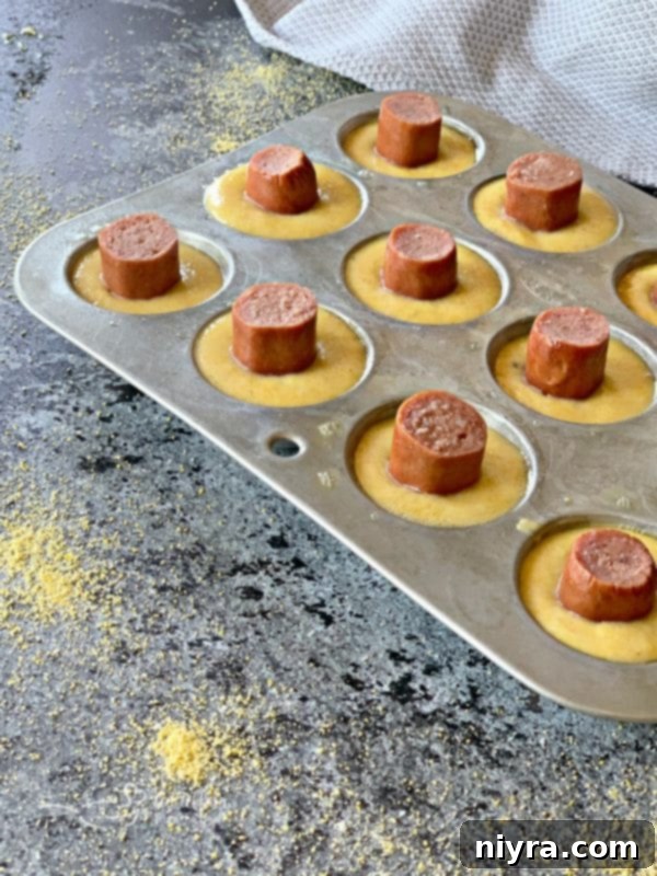 Hot dog pieces being placed into batter in a mini muffin tin for Corn Dog Mini Muffins
