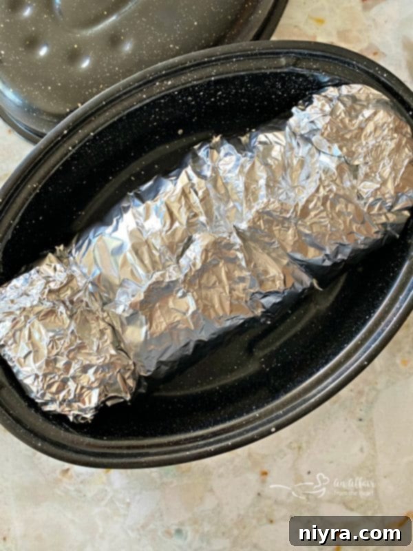 Pork roast tightly wrapped in aluminum foil, placed in a roasting pan, ready to be baked for hours in the oven.