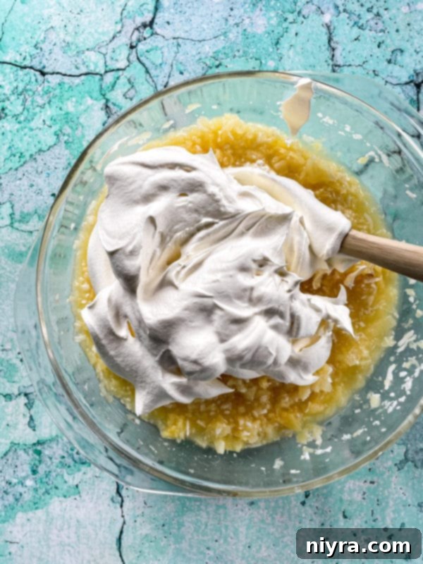 Thawed Cool Whip being gently folded into the pineapple and pudding mixture in a clear bowl.