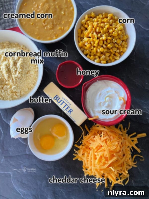 Golden Corn Bake 7 A flat lay of corn pie ingredients including creamed corn, corn muffin mix, butter, eggs, cheese, sour cream, and honey on a rustic grey surface.