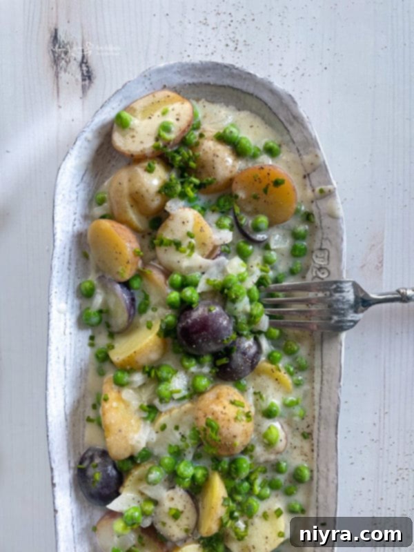 A white serving dish filled with cooked new potatoes and peas generously coated in Béchamel sauce.