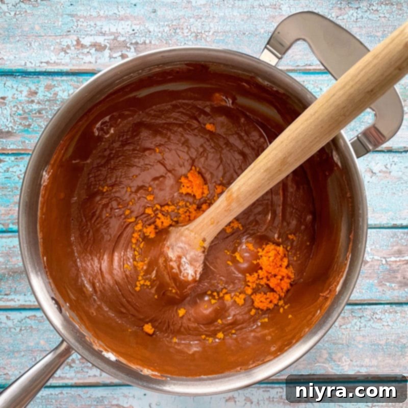 Orange zest being stirred into the melted fudge mixture in a saucepan with a wooden spoon, capturing the moment flavors infuse.