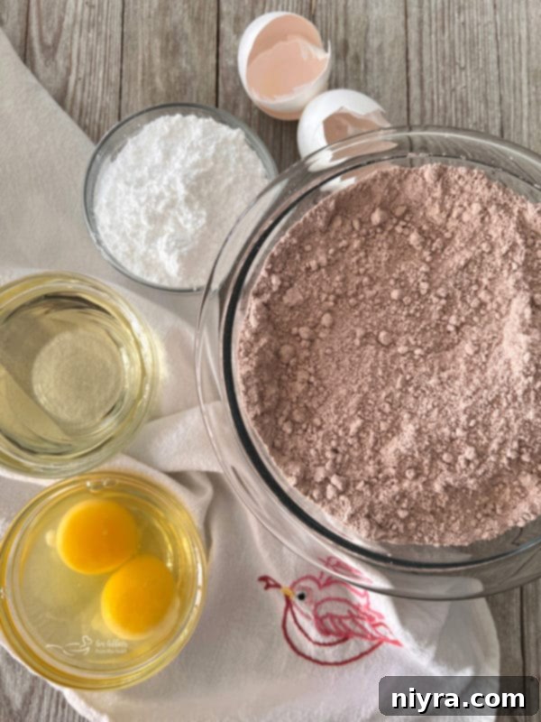 Ingredients for red velvet crinkle cookies: a large bowl of red velvet cake mix surrounded by smaller bowls containing vegetable oil, eggs, and powdered sugar.
