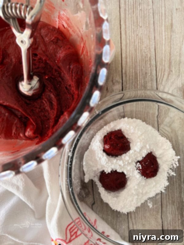 A bowl filled with vibrant red red velvet cake mix batter, next to a separate bowl overflowing with powdered sugar, ready for coating.