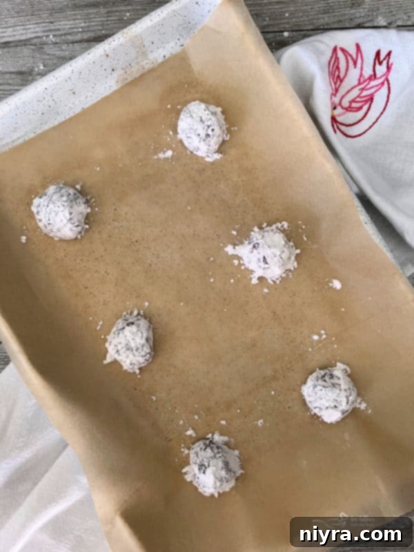 Multiple red velvet cookie dough balls, heavily coated in powdered sugar, neatly arranged on a parchment-lined baking sheet, ready for the oven.