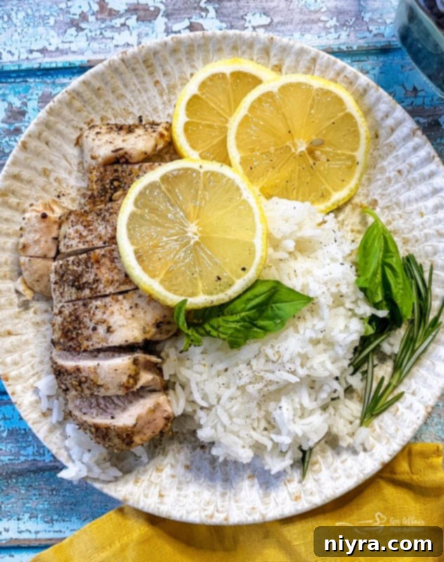 A plate featuring a perfectly cooked lemon pepper chicken breast, served alongside fluffy rice, a fresh lemon wedge, and sprigs of rosemary and basil for garnish.