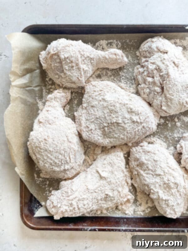 Chicken pieces draining on a wire rack over a baking sheet, with hot oil heating in a cast iron skillet.