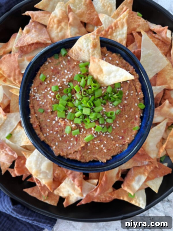 A vibrant blue bowl filled with Asian Bean Dip, garnished with fresh green onions, sits next to a jar of Paisley Farm Asian Bean Salad and a variety of colorful dipping vegetables.