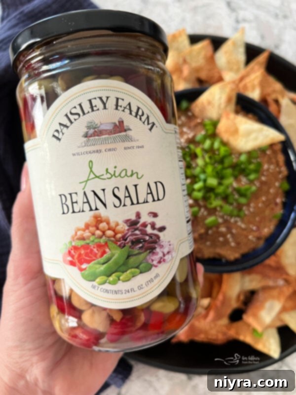 A glass jar of Paisley Farm Asian Bean Salad, showcasing a medley of colorful beans and vegetables, sits prominently on a wooden surface.