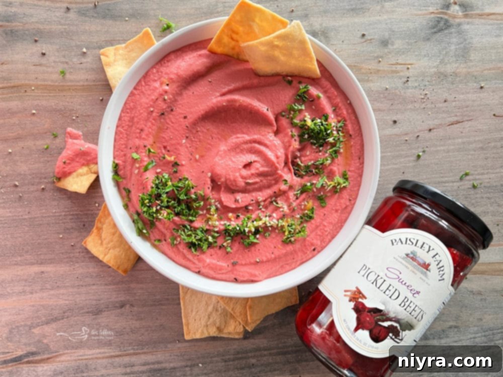 A bowl of vibrant pink pickled beet hummus, garnished with fresh parsley, sits beside a basket of pita chips and a jar of Paisley Farm Sweet Pickled Beets.
