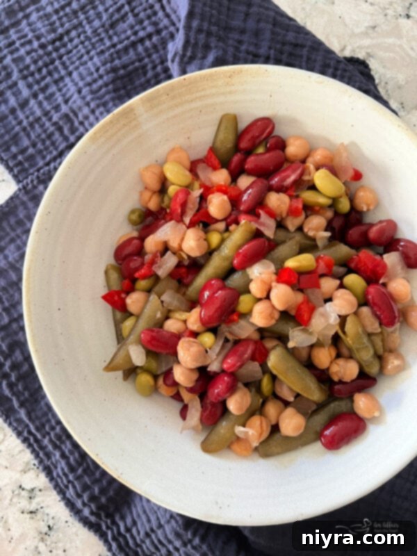A close-up of Paisley Farm Asian Bean Salad in a glass jar, showing the distinct texture and colors of snow peas, beans, and edamame, against a blurred background.