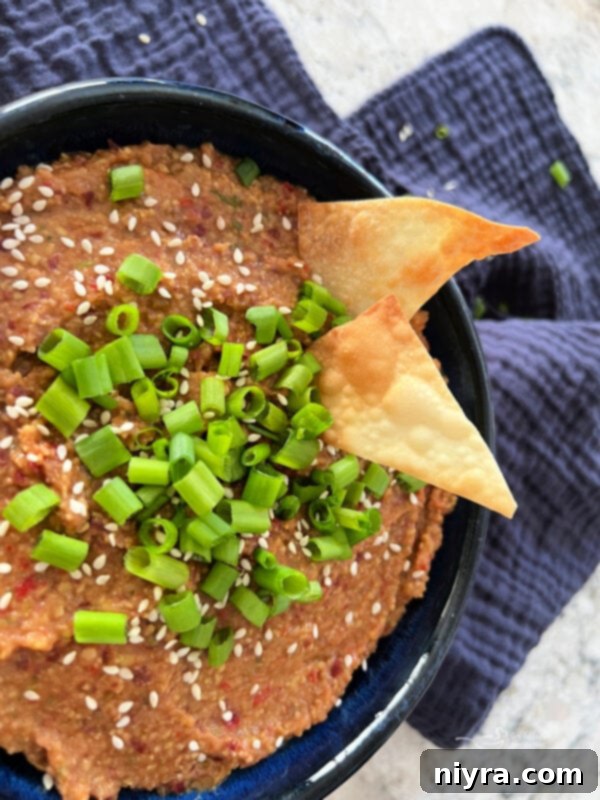 A bowl of creamy Asian Bean Dip, garnished with sliced green onions and toasted sesame seeds, is served with an assortment of fresh cucumber slices, carrot sticks, and bell pepper strips for dipping.