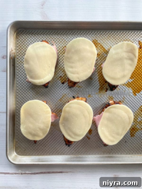 Smoky Mountain Chicken on a baking sheet, with ham and provolone cheese layered on top, ready for the oven.