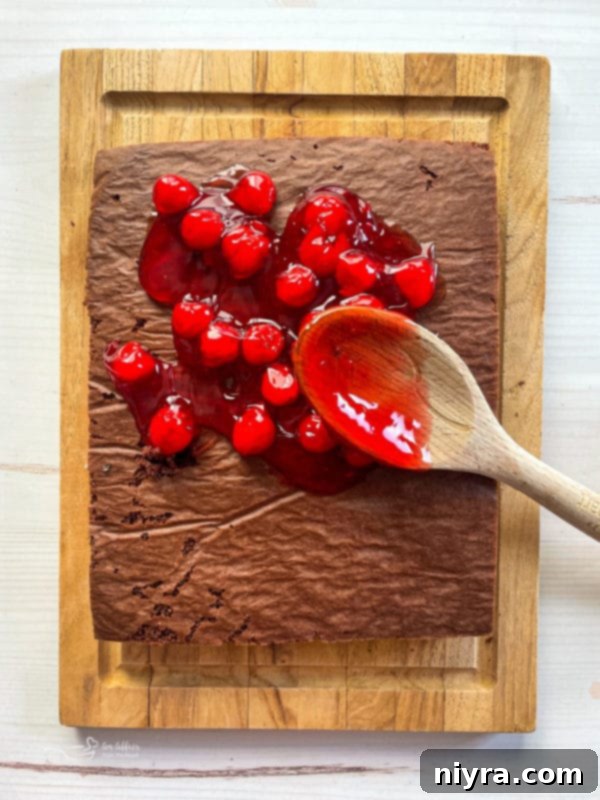 A spatula delicately spreading vibrant red cherry pie filling over a baked chocolate cake layer, preparing for the next layer of this delicious Black Forest Cake.