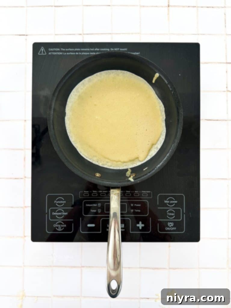 Instructional image showing crepe batter being poured into a hot skillet, with a thin layer spreading across the pan.