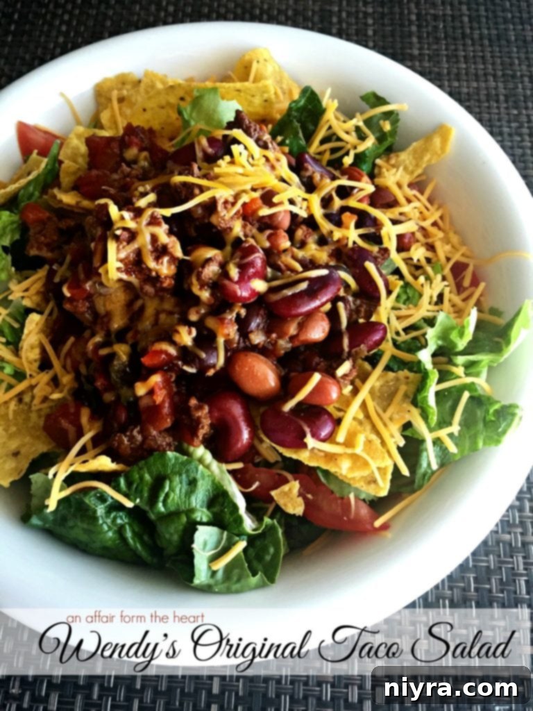 A vibrant Copycat Wendy's Original Taco Salad with a generous serving of chili, fresh lettuce, cheese, and crunchy tortilla chips in a white bowl