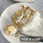 A fork with a bite of the COCONUT CREAM POKE CAKE WITH COCONUT WHIPPED CREAM FROSTING next to the slice of cake on a white plate