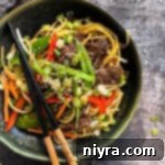 Close up of Chops sticks atop the Beef Lo Mein in a dark green bowl.